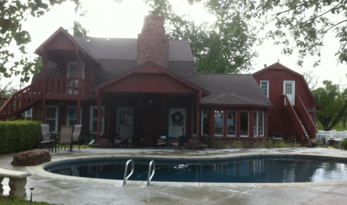 A red two-story house with a pool in the foreground, surrounded by trees and outdoor seating.