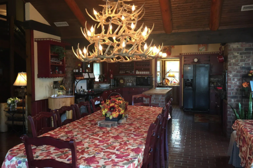 Cozy kitchen with a large dining table, rustic decor, and an antler chandelier, featuring warm colors and a homey atmosphere.