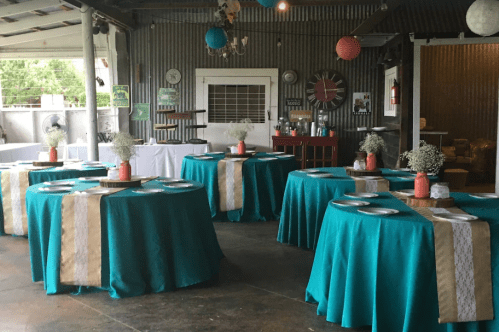 A rustic venue with teal tablecloths, centerpieces of flowers, and decorative elements hanging from the ceiling.