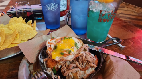 A sizzling skillet with pulled pork, a fried egg, and green onions, accompanied by chips and colorful drinks.