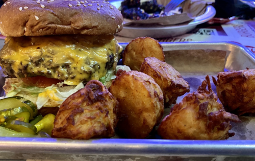 A sesame seed bun burger with melted cheese, lettuce, and tomato, served with crispy potato bites on a metal tray.
