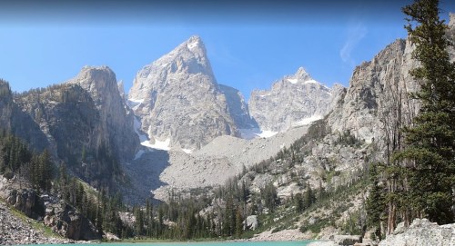 A panoramic view of rugged mountains with snow-capped peaks and a turquoise lake surrounded by trees under a clear blue sky.