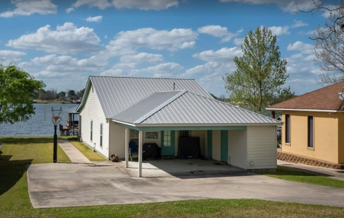 A lakeside house with a metal roof, surrounded by green grass and trees, under a partly cloudy sky.