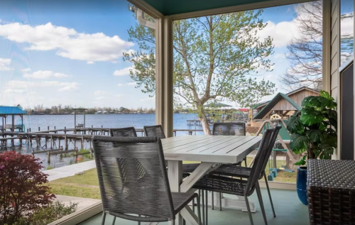 A cozy patio with a table and chairs, overlooking a serene lake and dock under a blue sky with fluffy clouds.