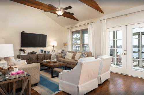 Cozy living room with a ceiling fan, large windows, and a view of the water, featuring comfortable seating and a TV.