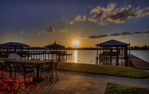 Sunset over a lake with docks and a patio area, featuring tables and chairs, surrounded by clouds and vibrant colors.