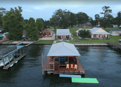 Aerial view of a lakeside dock with chairs, surrounded by houses and green trees along the water's edge.