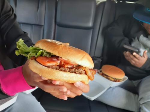 A person holds a large burger with lettuce, tomato, and bacon, while another person sits in the background using a phone.