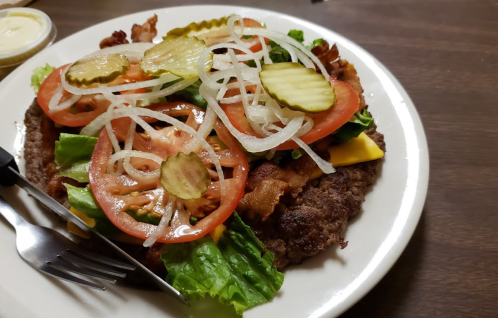 A plate with a large beef patty topped with lettuce, tomato, pickles, onions, and cheese, served with a fork and knife.