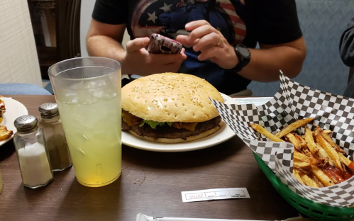 A large burger with lettuce sits on a table next to a basket of fries and a glass of drink. A person is using a phone.