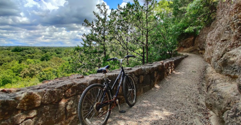A mountain bike rests on a gravel path beside a stone wall, surrounded by lush greenery and a cloudy sky.