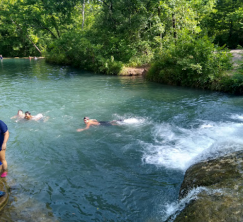 Two people swim in a clear, greenish-blue river surrounded by lush greenery, with a small waterfall in the foreground.