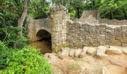 A stone bridge arches over a small stream, surrounded by lush greenery and remnants of a historic structure.