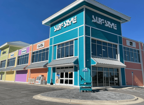 Colorful storefronts of a shopping center with "Surf Style" prominently displayed on the main building. Clear blue sky above.