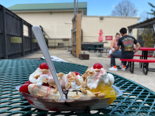 A colorful sundae with whipped cream, cherries, and nuts sits on a green table, with people in the background.