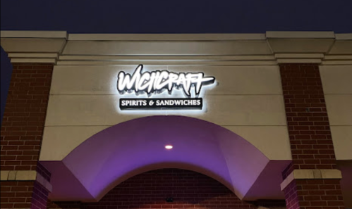 Sign for "Witchcraft Spirits & Sandwiches" illuminated on a brick building at night.