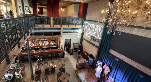 A spacious restaurant interior with a bar, dining tables, and a stage, featuring a chandelier and rustic decor.