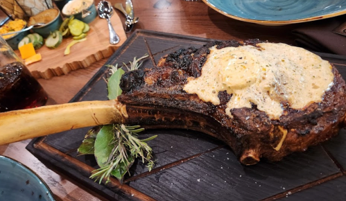 A large, charred tomahawk steak topped with a creamy sauce, garnished with herbs, served on a wooden platter.
