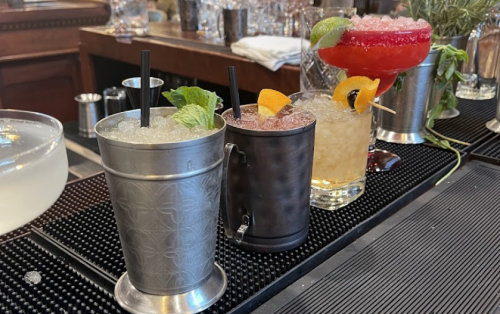 Three colorful cocktails with garnishes on a bar counter, featuring mint, citrus, and decorative elements.