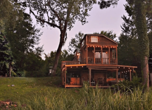 A charming wooden house with string lights, surrounded by greenery and trees, set against a twilight sky.