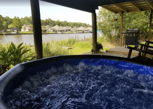 A view from a deck overlooking a river, featuring a hot tub and nearby greenery, with houses in the background.