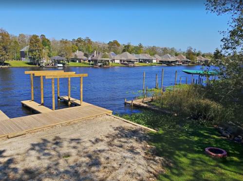 A serene river view with a wooden dock, houses along the shore, and lush greenery under a clear blue sky.