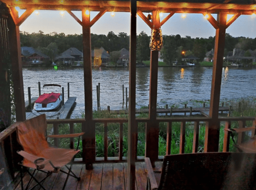 View from a porch overlooking a river with boats, surrounded by greenery and illuminated by string lights at dusk.