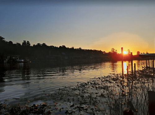 Sunset over a calm river, with reflections on the water and silhouettes of trees and docks in the background.