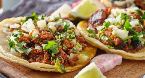 Close-up of tacos topped with chopped onions, cilantro, and lime wedges on a wooden board.