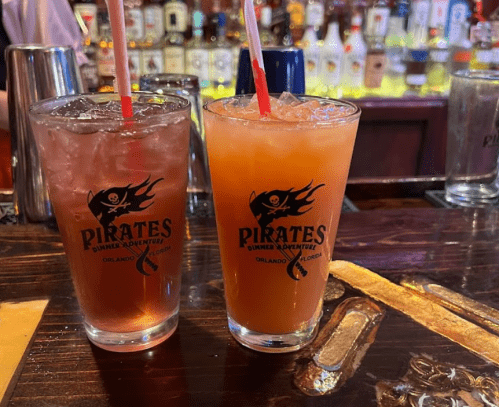 Two colorful cocktails in glasses with a pirate logo, sitting on a bar with various bottles in the background.