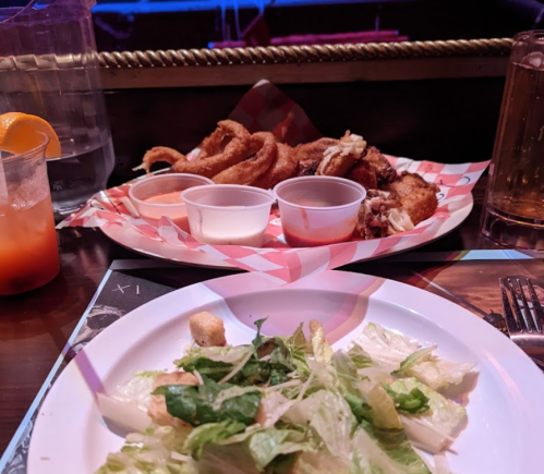 A table setting with a plate of fried food, dipping sauces, a salad, and drinks in a dimly lit venue.