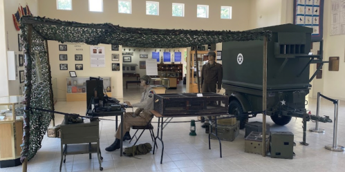 A military exhibit featuring a soldier at a desk, a camouflaged tent, and a vintage military vehicle in a museum setting.