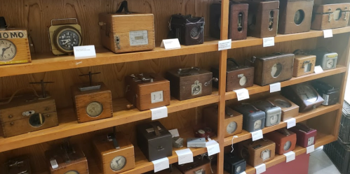 A display of various vintage clocks and timepieces on wooden shelves, each with unique designs and labels.