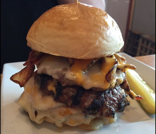 A stacked burger with two patties, melted cheese, bacon, and a toasted bun, served with a pickle on the side.