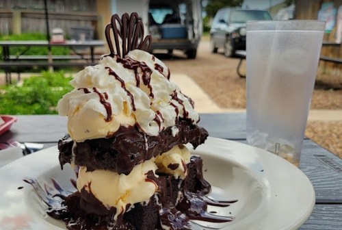 A towering dessert of chocolate cake, ice cream, whipped cream, and chocolate drizzle, served on a plate with a drink nearby.