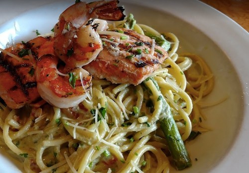 A plate of pasta with grilled shrimp and salmon, topped with asparagus and herbs in a creamy sauce.