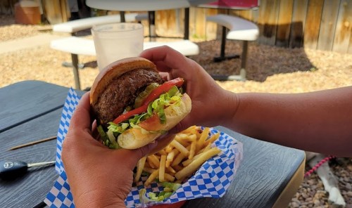 A person holds a burger with lettuce, tomato, and pickles, with a side of fries on a table outdoors.