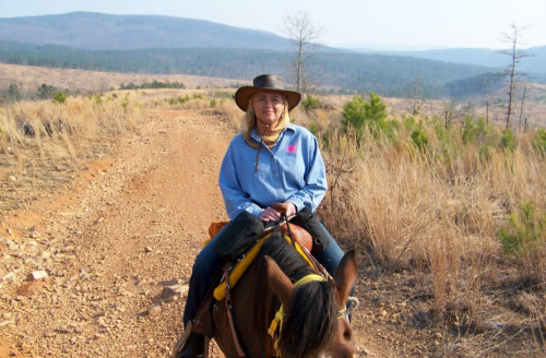 A woman in a hat sits on a horse on a dirt trail surrounded by dry grass and distant hills.