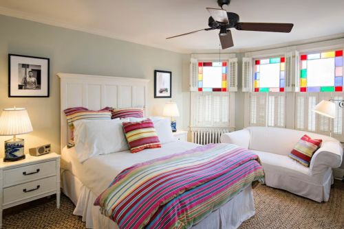 Bright bedroom with a striped bedspread, white furniture, a fan, and colorful stained glass windows. Cozy and inviting atmosphere.