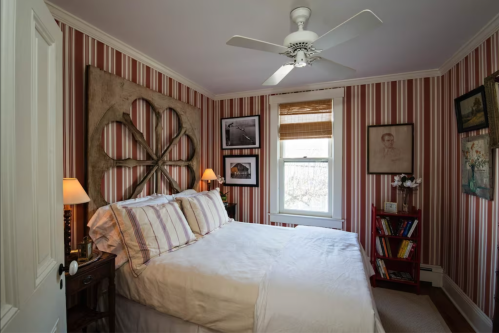 Cozy bedroom with striped walls, a large bed, ceiling fan, and framed artwork, featuring a warm, inviting atmosphere.
