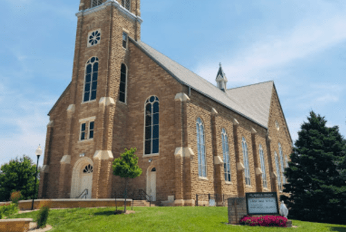 A large stone church with tall windows and a steeple, surrounded by greenery and a clear blue sky.