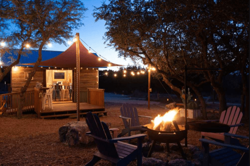 Cozy cabin at dusk with string lights, a fire pit, and Adirondack chairs in a serene outdoor setting.