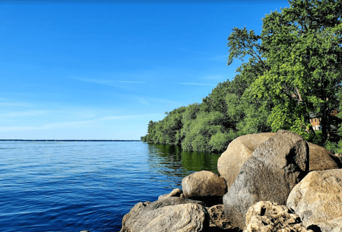 A serene lakeside view with clear blue water, rocky shore, and lush green trees under a bright blue sky.