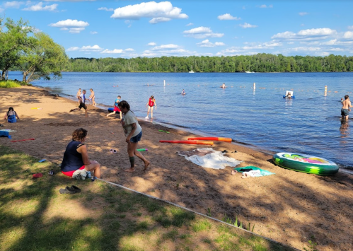 People enjoying a sunny day at the beach, swimming and relaxing by the water with trees in the background.