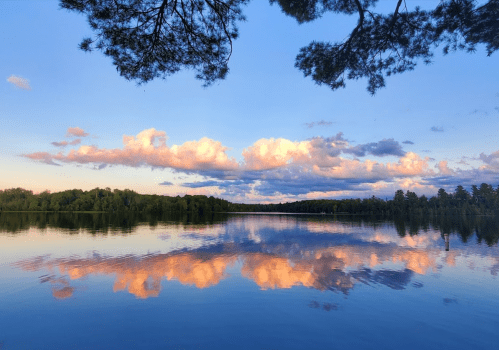 A serene lake at sunset, reflecting colorful clouds and surrounded by lush greenery under a clear blue sky.