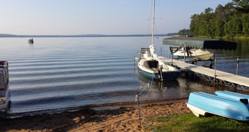 A calm lake scene with boats docked, a sandy shore, and trees lining the background under a clear blue sky.