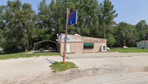 A small, single-story building with a sign reading "Aeroplane Inn," surrounded by trees and a gravel parking area.