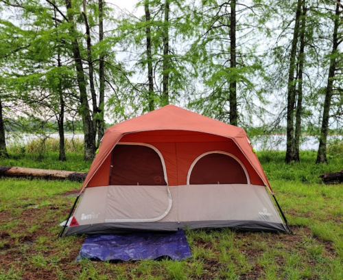 A brown and beige tent set up on green grass near a lake, surrounded by trees.