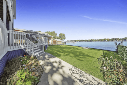 A serene lakeside view featuring a grassy area, steps leading to a house, and clear blue skies.