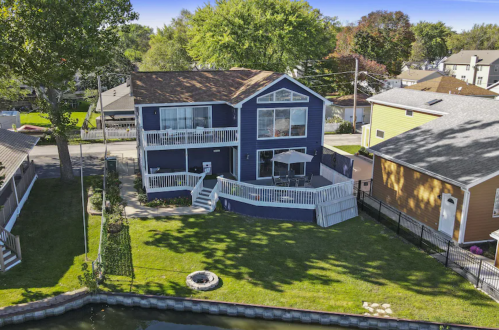 Aerial view of a blue house with a deck, surrounded by greenery and a canal, with neighboring homes in the background.